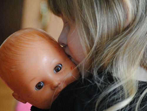 Menina de costas segurando uma boneca de brinquedo e a beijando na cabeça.