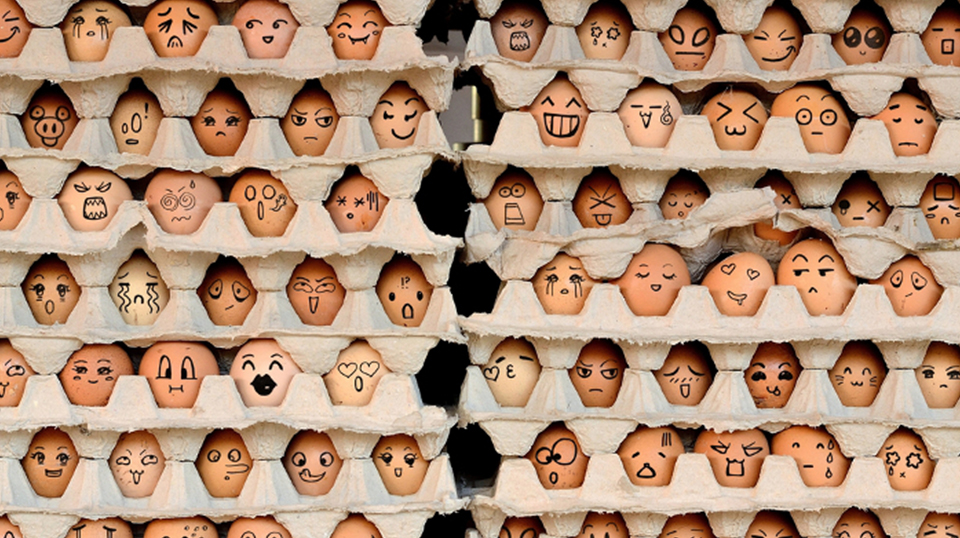 Caixas de ovos dispostas uma sob as outras. Nos ovos estão desenhadas diversas rostos e expressões diferentes.