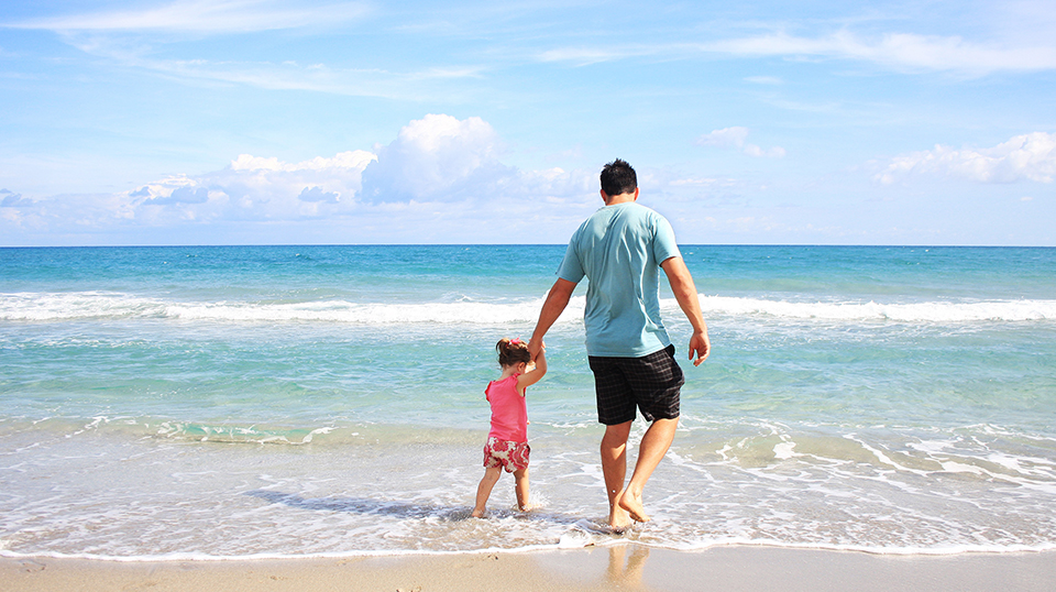 Pai segurando a mão da filha pequena na beira do mar, ambos estão de costas.