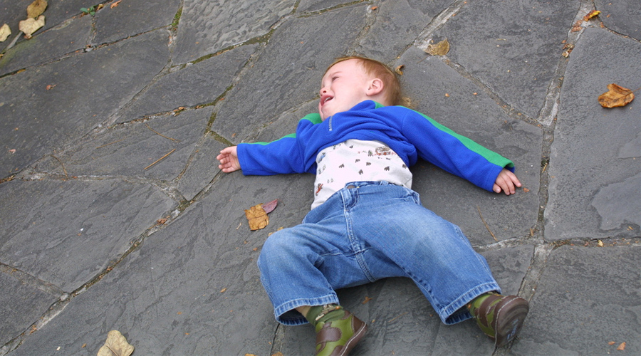 Menino de calça jeans e jaqueta azul atirado na calçada de pedra fazendo birra.