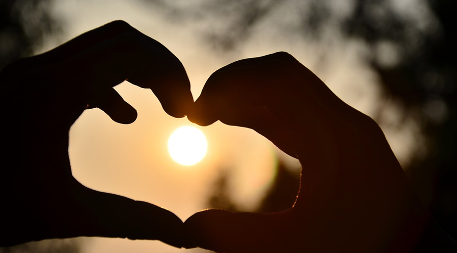 Duas mãos formando um coração com o sol ao fundo.
