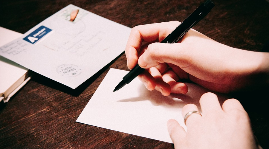 Mão segurando uma caneta e apoiada sob uma mesa e uma folha de papel em branco.