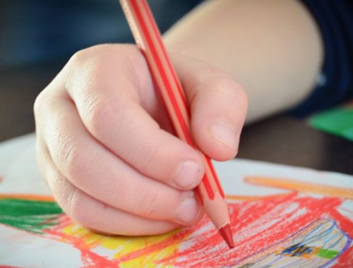 Mão de uma criança segurando um lápis de cor vermelho e pintando um desenho.