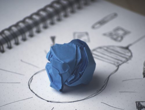 Papel azul amassado feito bolinha em cima de um papel branco com um desenho de uma lâmpada.