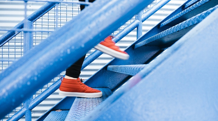Foto lateral de pés calçados com tênis vermelho subindo uma escada azul.