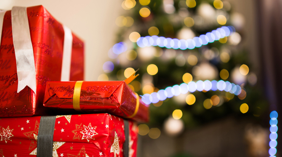 Foto de três presentes de pacotes vermelhos empilhados e, ao fundo, uma árvore de Natal com luzes azuis.